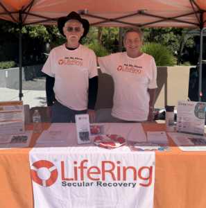 Two smiling men in LifeRing shirts stand at an outdoor information booth under an orange canopy, with brochures on the table in front of them.